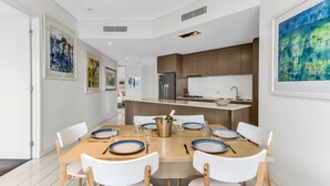 A modern dining area with a central island, white chairs, and a kitchen in the background. - Tropical Park View Retreat – 3BR Bali Resort in Kingscliff (1210) (Tweed)