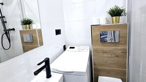 A modern bathroom with a white sink, a black faucet, a mirror, a towel dispenser, a shower area, and a wooden panel with a picture. - Studio Bajka - 365PAM (Mielno)