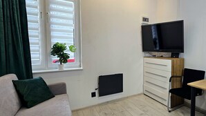 A room with a sofa, a TV, a small table, a plant, and a window with blinds. - Studio Bajka - 365PAM (Mielno)