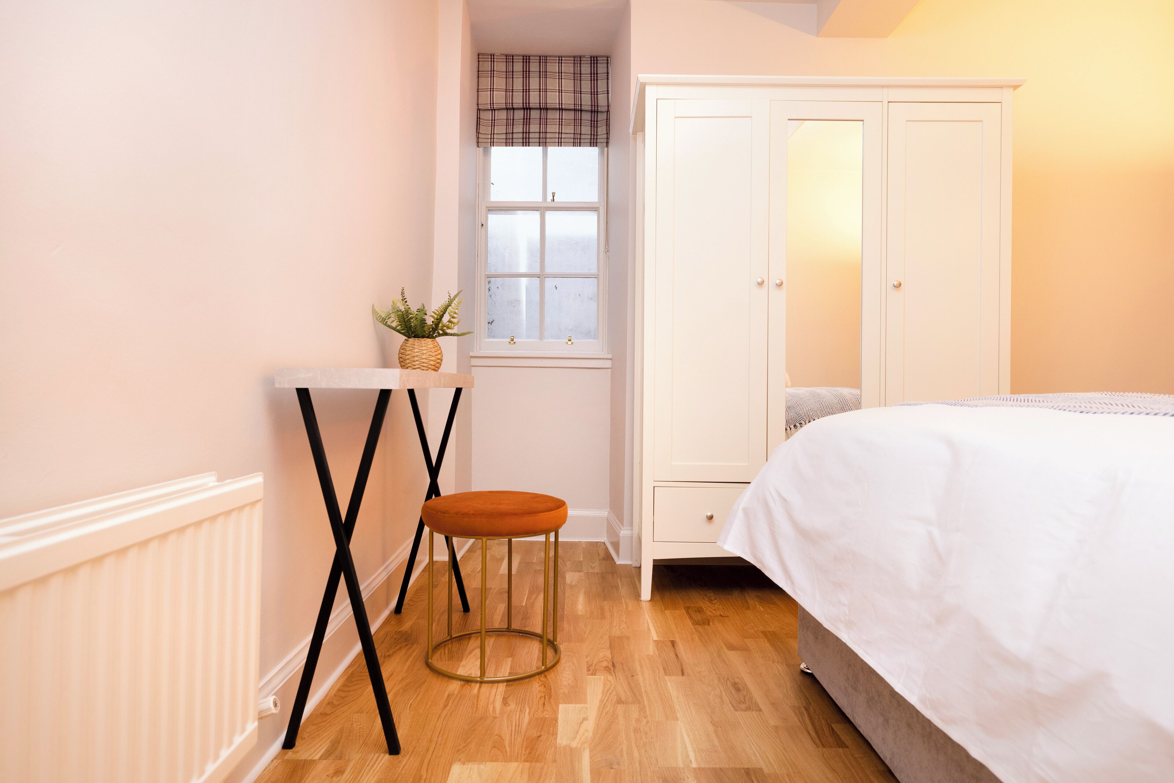 A bedroom with a bed, a white wardrobe, a window with a plaid valance, a small stool, and a plant on a table.