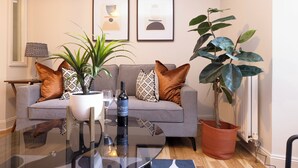 Una sala de estar moderna con un sofá gris, cojines estampados, una mesa de centro de vidrio, una planta en maceta y cuadros en la pared.