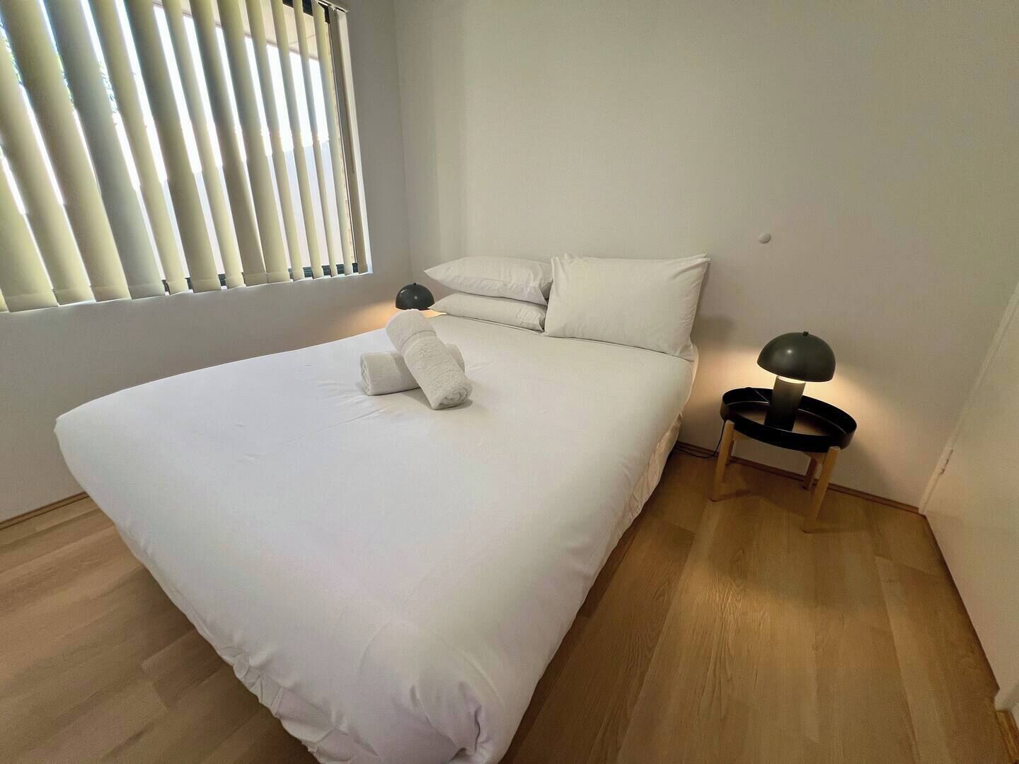 Una cama bien hecha con sábanas blancas y dos toallas enrolladas, una lámpara de noche y una ventana con persianas.