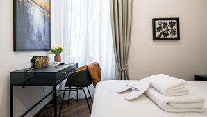 Een hotelkamer met een bed, een bureau met een stoel, een kleine tafel met een plant, een lijst met kunstwerk en een raam met gordijnen.