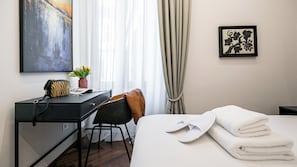 A hotel room with a bed, a desk with a chair, a small table with a plant, a framed artwork, and a window with curtains.