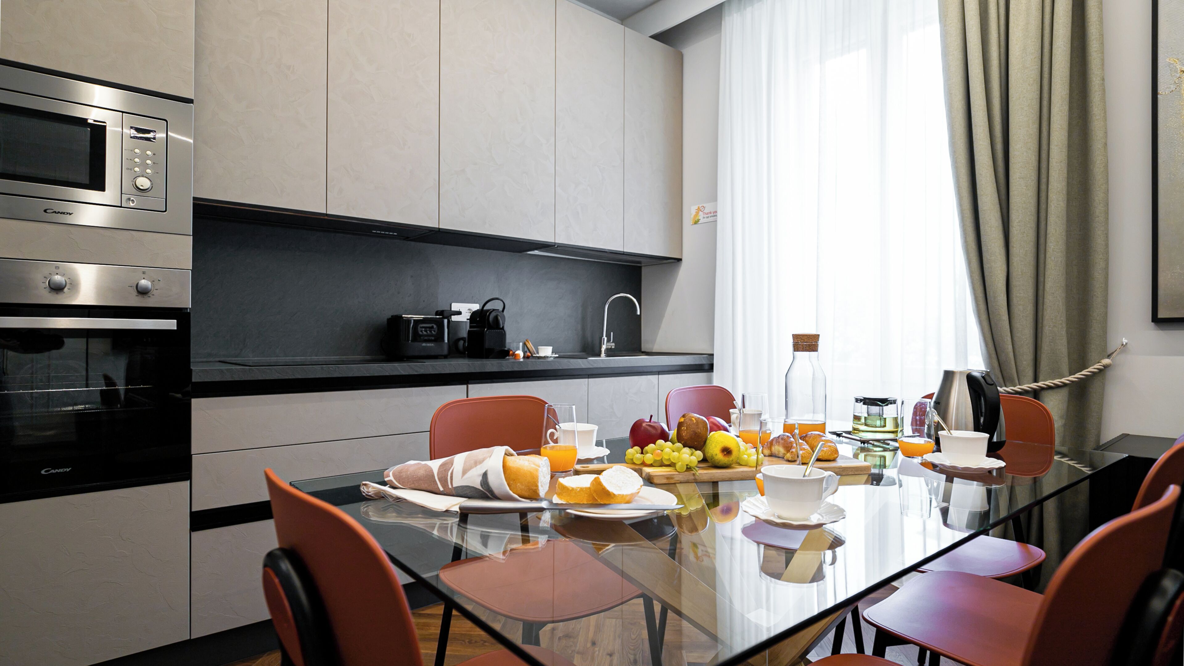 A modern kitchen with a microwave, oven, and coffee maker, a dining table with chairs, and breakfast items.