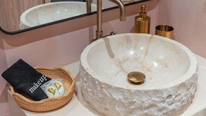 Une salle de bain avec un lavabo rond en marbre, un robinet doré, un distributeur de savon doré et un panier en osier contenant une serviette noire et une trousse de maquillage.