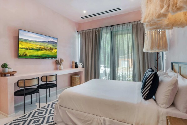Un dormitorio con cama, escritorio, dos sillas, televisor y ventana con cortinas.