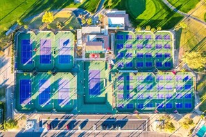 An aerial view of a sports complex with multiple tennis courts, a parking area, and surrounding greenery.
