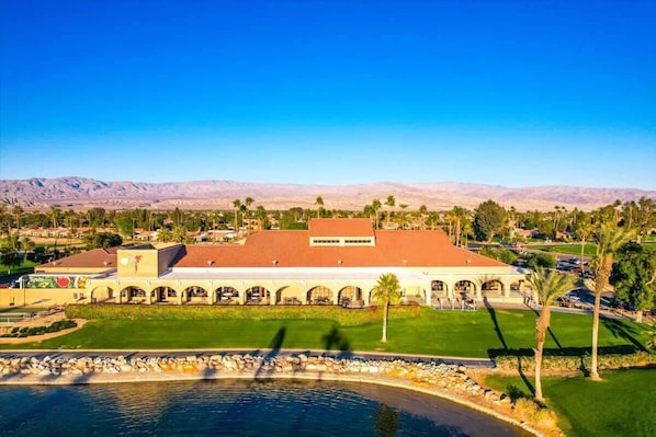 A hotel with a large building featuring arched walkways and a red roof, situated near a body of water with a mountainous backdrop.