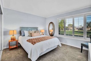 A bedroom with a bed, two bedside lamps, a large mirror, and a window with blinds. - Fairway Retreat | 2BR Resorter (Palm Desert)
