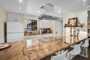 A modern kitchen with white cabinets, a marble island, and bar stools. - Fairway Retreat | 2BR Resorter (Palm Desert)
