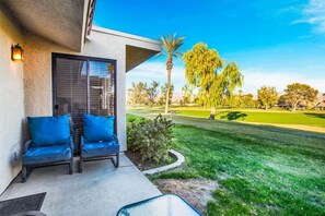 A modern building with a covered patio area featuring two blue cushioned chairs and a glass table. - Fairway Retreat | 2BR Resorter (Palm Desert)