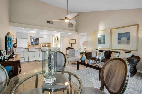A spacious living room with a glass coffee table, a sofa, and a dining area with chairs. - Fairway Retreat | 2BR Resorter (Palm Desert)