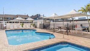 A swimming pool with a tiled edge, surrounded by lounge chairs and umbrellas, with buildings in the background. - Absolute Beachfront Apartment 4T (Lennox Head)