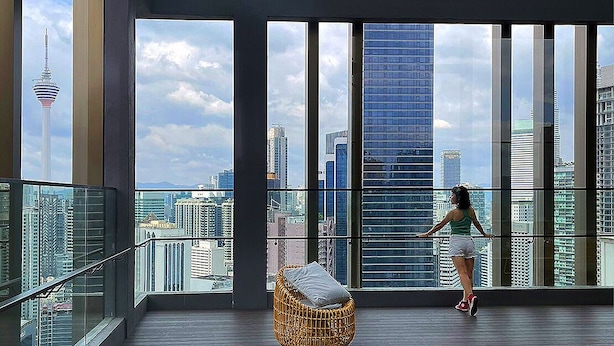 Un balcon spacieux offrant une vue sur les toits de la ville, une chaise en osier et une personne se tenant près de la balustrade.