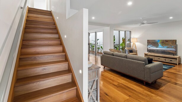 A modern living room with a wooden staircase, a grey sofa, a flat-screen TV, and a ceiling fan. - Nautilus - Lennox Head (Lennox Head)