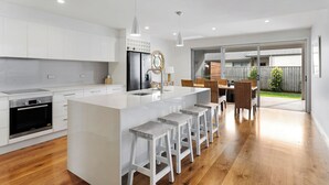 A modern kitchen with white cabinetry, a central island with bar stools, a built-in oven, and a dining area with a view of the backyard. - Nautilus - Lennox Head (Lennox Head)