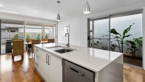A modern kitchen with a white countertop, stainless steel dishwasher, and a dining area with wicker chairs. - Nautilus - Lennox Head (Lennox Head)