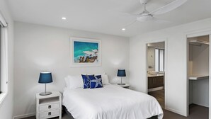 A modern bedroom with a large bed, two bedside tables with lamps, a ceiling fan, a painting, and a bathroom visible through an open door. - Nautilus - Lennox Head (Lennox Head)