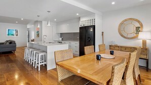 A modern kitchen with white cabinets, a black refrigerator, and a dining area with a wooden table and wicker chairs. - Nautilus - Lennox Head (Lennox Head)