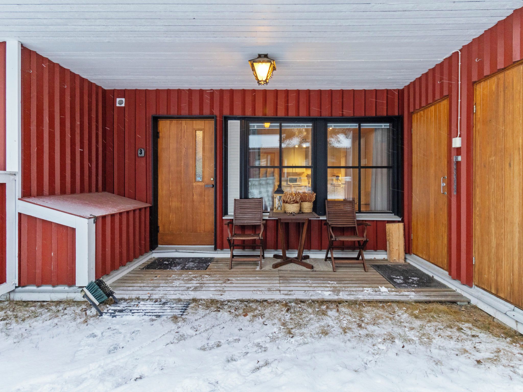 Een overdekte veranda met een houten tafel en twee stoelen, een bankje en een deur met een nummer.