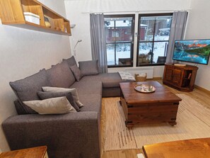 A living room with a sectional sofa, a wooden coffee table, a television, and a window with curtains.