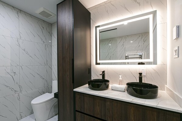 Un baño moderno con baldosas de mármol, un mueble de tocador de madera oscura, dos lavamanos negros y un espejo grande con iluminación integrada.