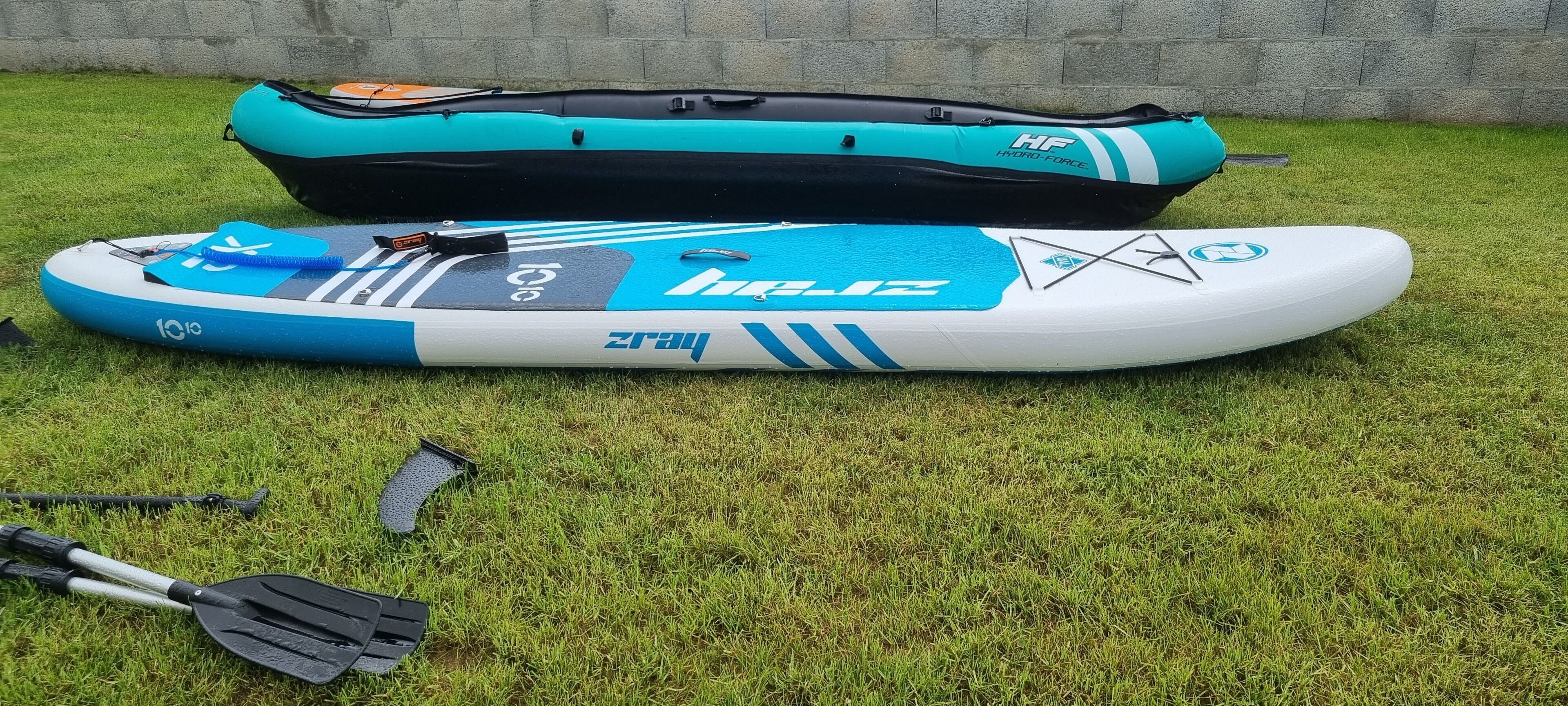 A blue and white inflatable raft with a paddle and a black and teal inflatable bag on a grassy lawn.