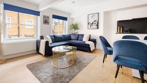 A modern living room with a dark blue sofa, a round glass coffee table, and matching blue chairs. - LONDONERS│Sherlock's Home  (London)