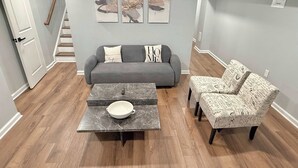 Ein Wohnzimmer mit grauem Sofa, einem Marmortisch, zwei gemusterten Sesseln und einer Treppe.