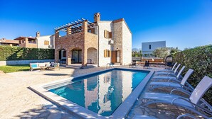 Un área de piscina con sillones de descanso, una piscina de piedra y un edificio de ladrillo con un porche cubierto.