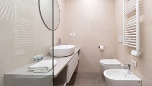 Un baño moderno con un lavamanos ovalado grande, un inodoro y un bidé. Hay un espejo sobre el lavamanos y un perchero con toallas.