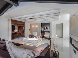 A modern living room with a sofa, a coffee table, and a TV. There is a kitchen area with a bar and a dining table.