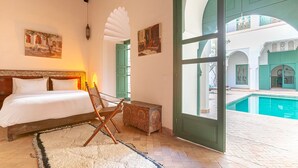 A bedroom with a bed, a chair, a small table, a large window, and a view of an outdoor pool area. - Dar Salam, swimming pool, rooftop, staff, Medina (Marrakech)