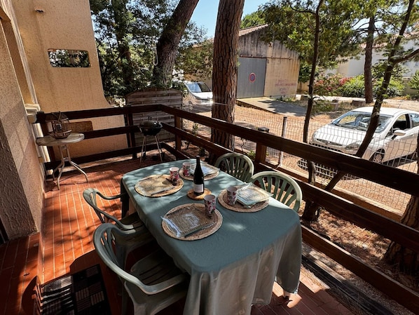 Un patio exterior con una mesa lista para dos personas, rodeado de sillas, una parrilla y vistas a coches aparcados y árboles.
