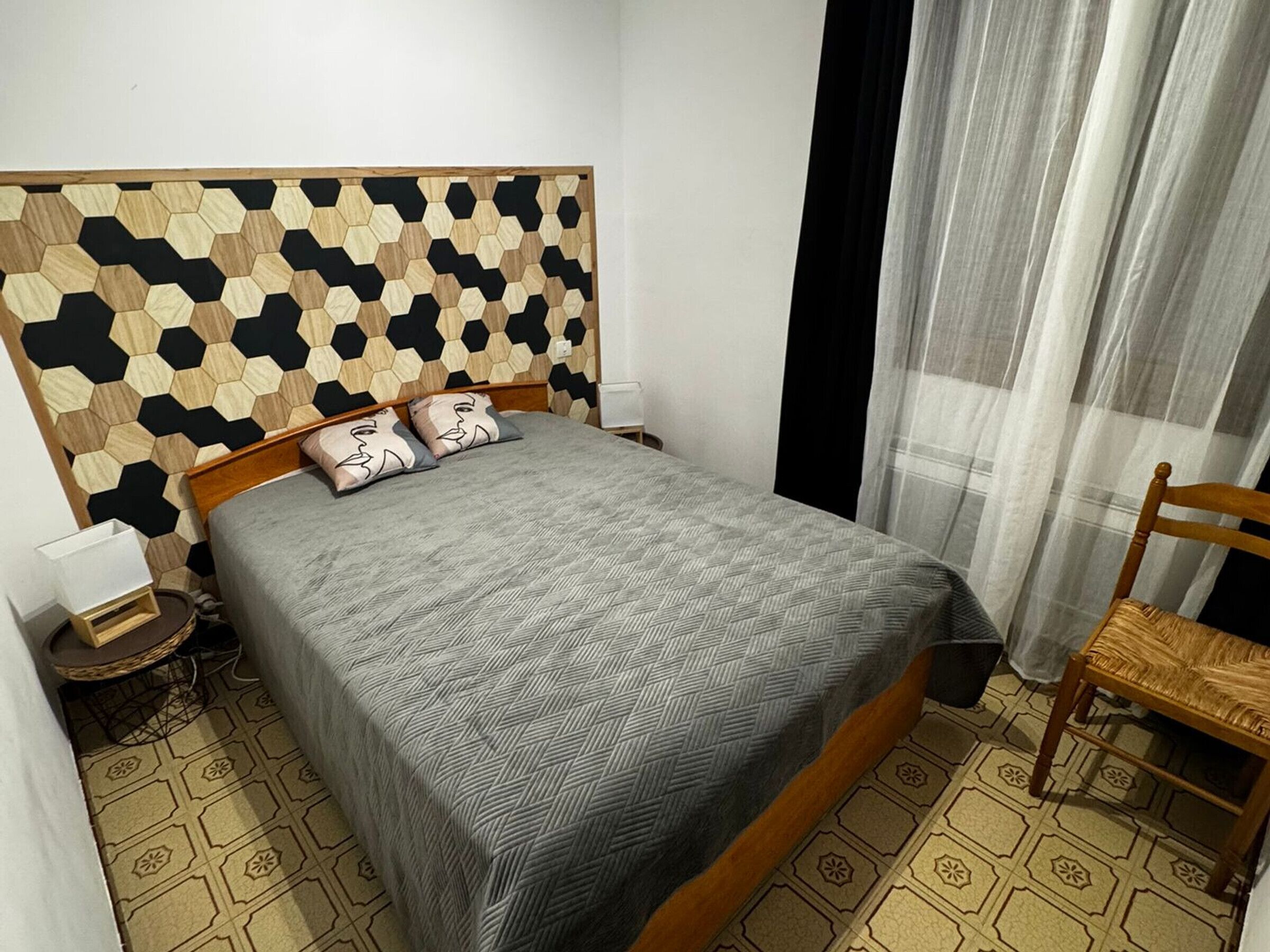 Une chambre à coucher avec une tête de lit en bois, un lit recouvert d’une literie à motifs, une table de chevet avec une lampe, une chaise et des carreaux de sol à motifs.