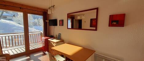A room with a wooden table and chairs, a large mirror, and a TV. There is a balcony with a view of snow-covered trees.