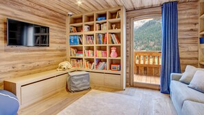 L’intérieur chaleureux d’un chalet avec une bibliothèque en bois, un téléviseur, un canapé et une vue sur une montagne boisée.