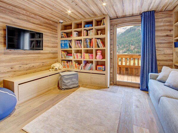 A cozy cabin interior with a wooden bookshelf, a television, a sofa, and a view of a forested mountain. - 5★ chalet, 15 people, indoor pool, mountain views, 7 bedrooms, terrace, pets welcome (La Clusaz)