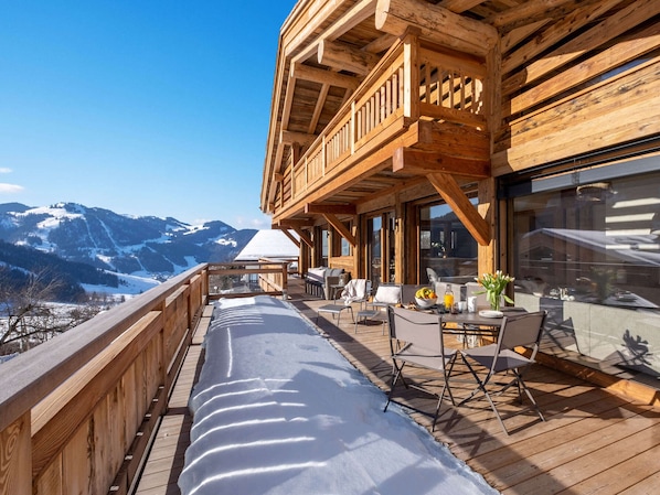 A wooden deck with outdoor seating, a view of snow-covered mountains, and a large window.