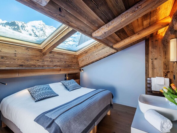 A bedroom with a skylight, wooden beams, a bed with patterned pillows, a bedside table with a towel, and a view of snow-capped mountains. - 5★ Chalet for 15 people, sauna, 300m from slopes (La Clusaz)