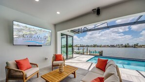 Un moderno espacio de sala de estar con piscina, mobiliario de mimbre y un televisor grande que muestra 'CoralVilla Villa & Boat Rental'.