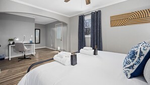 A modern hotel room with a wooden floor, a desk with a chair, a bed with white linens, a ceiling fan, and a window with curtains.