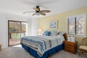 A bedroom with a bed, a ceiling fan, a nightstand, a chair, a window with blinds, and a door leading to a balcony.