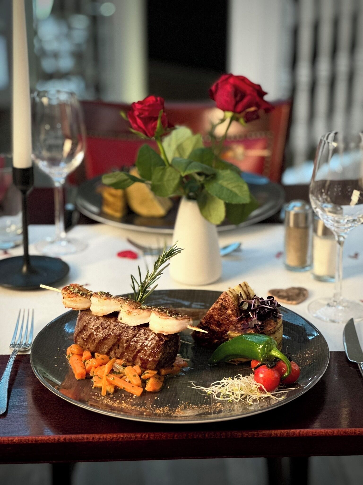 A dining table set with a plate of food, a vase with red roses, and wine glasses.