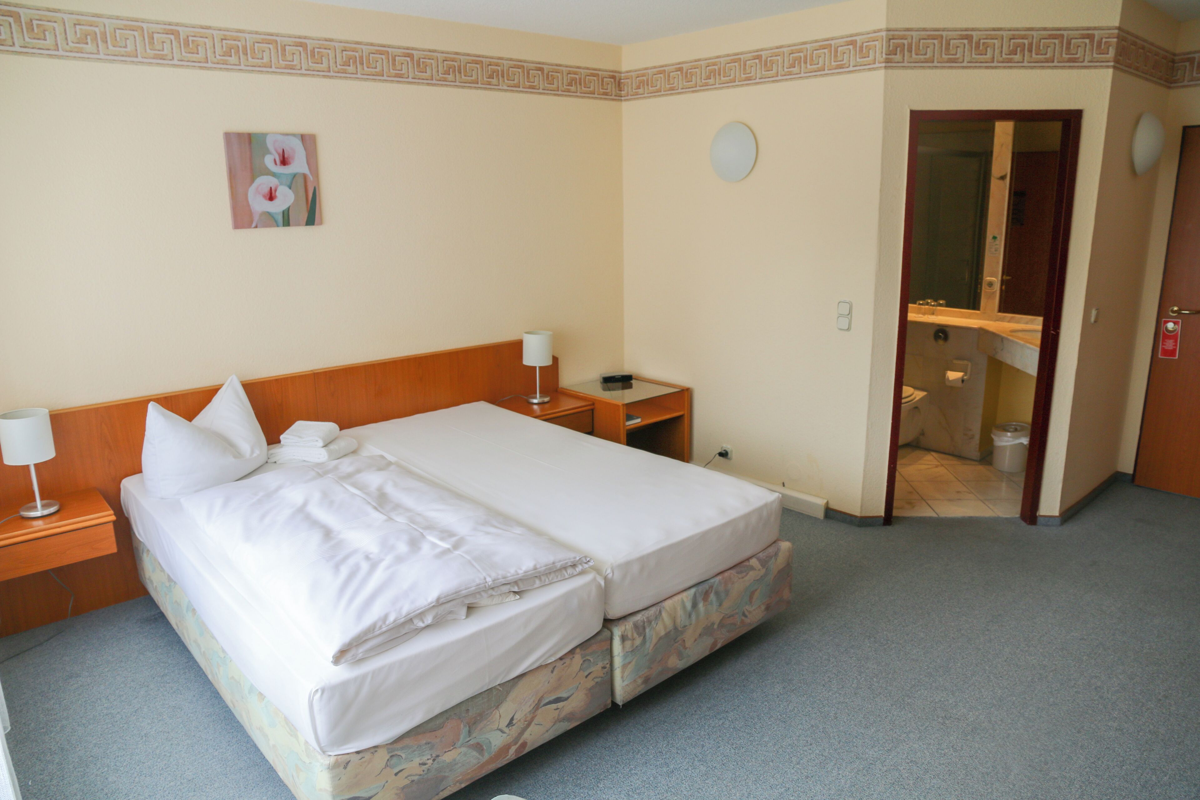 Een tweepersoonsbed met witte lakens, een nachtkastje met een lamp, een klein tafeltje met een telefoon, en een badkamer zichtbaar door een geopende deur.