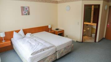 Een tweepersoonsbed met witte lakens, een nachtkastje met een lamp, een klein tafeltje met een telefoon, en een badkamer zichtbaar door een geopende deur.