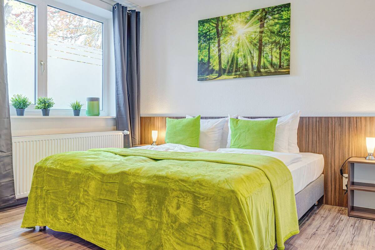 A hotel room with a bed, a window with curtains, a radiator, a bedside table with a lamp, and a painting of a forest.