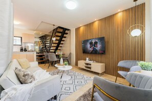 A modern living room with a sofa, armchairs, a TV, and a staircase leading to a kitchen.