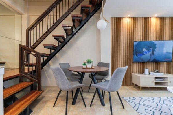 A modern living room with a wooden staircase, a round dining table with four chairs, a flat-screen TV mounted on a wooden wall, and a small white cabinet.